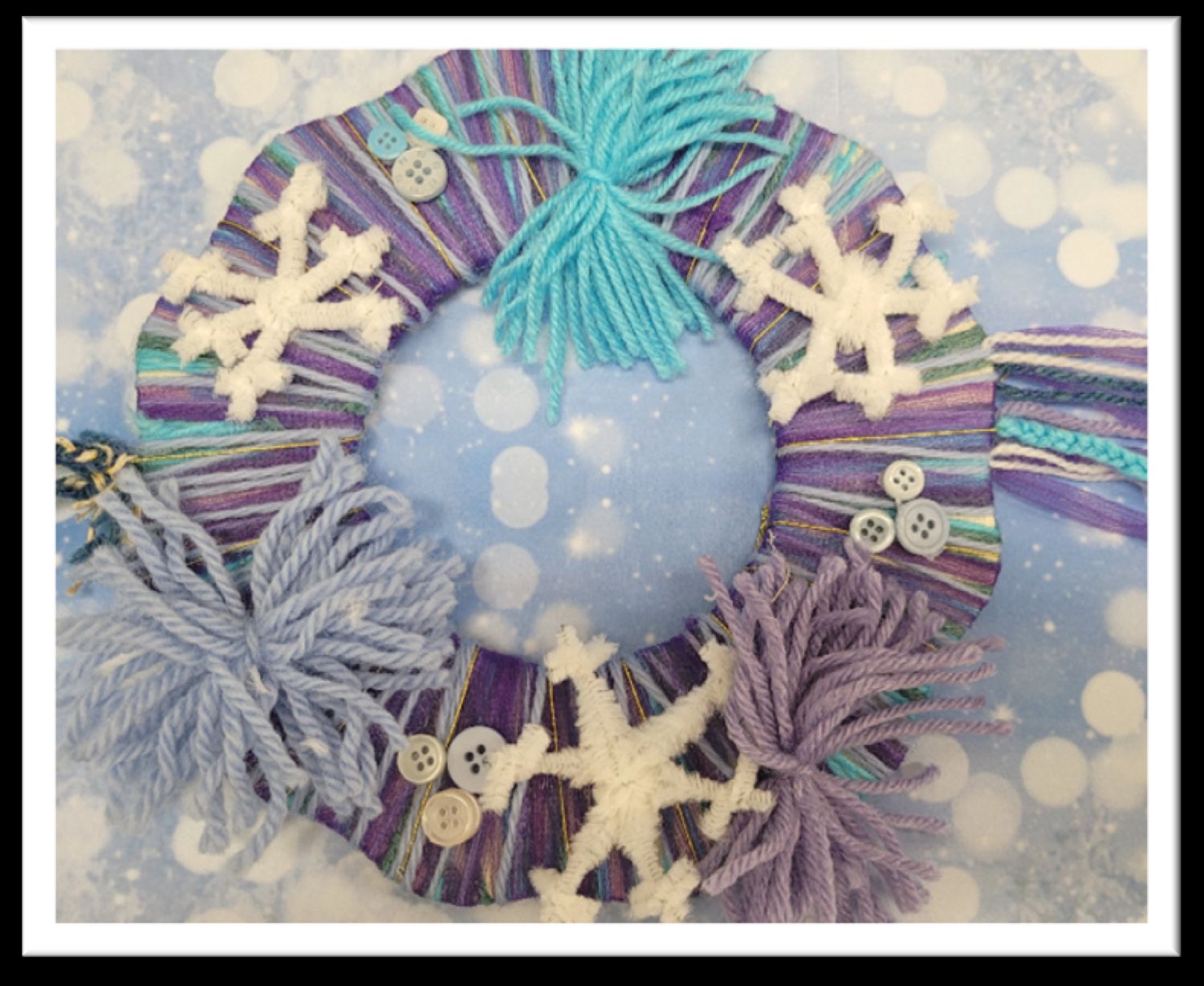 A handmade circular wreath is wrapped tightly with purple, blue, and teal yarn, decorated with white pipe cleaner snowflakes, multi-colored buttons, and fluffy yarn tassels against a snowy background.